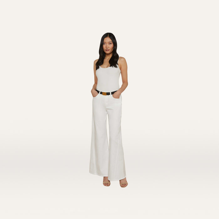 A woman stands against a plain white background wearing a white tank top, the Beau Wide-Leg Jean in high rise stretch denim, a black belt, and beige heels, with her hands in her pockets.