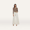 A woman stands against a plain white background wearing a brown collared long-sleeve shirt, the Beau Wide-Leg Jean in high-rise stretch cotton twill in white, a white belt with a gold buckle, and beige heels.