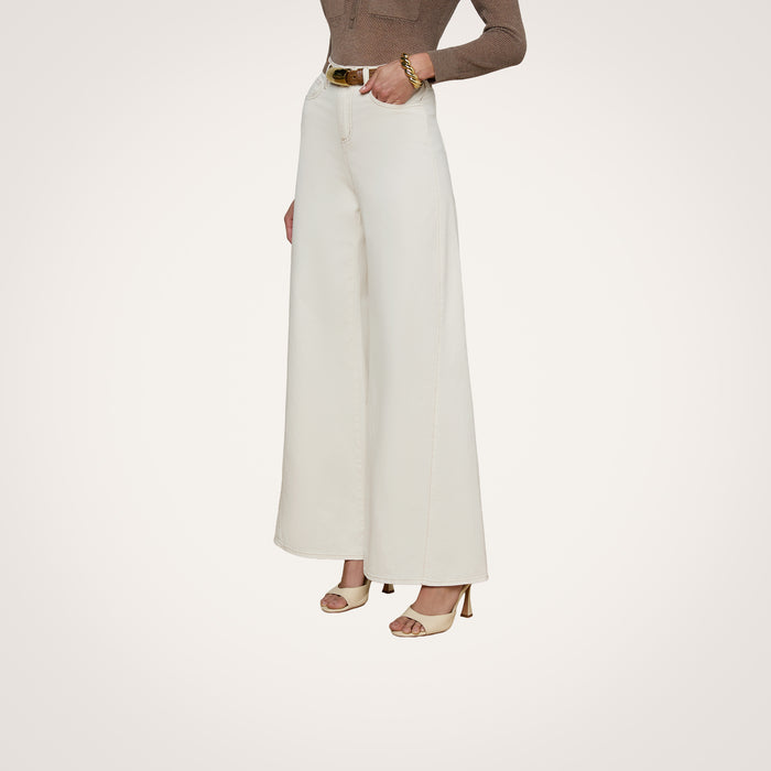 A person stands against a plain light background wearing the Beau Wide-Leg Jean—a high-rise, wide-leg style in white stretch cotton twill. Only the lower half of their body is visible, styled with beige heeled sandals, a belt, and a bracelet.