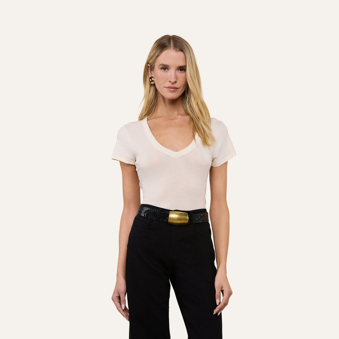 A woman with long blonde hair wears the Becca Cotton V-Neck Tee, a vintage off-white top made from 100 cotton, paired with black pants and a black belt with a gold buckle, standing against a plain light background.