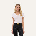 A woman with long blonde hair wears the Becca Cotton V-Neck Tee—crafted from 100 cotton in white—paired with black leather pants featuring a large gold buckle and gold jewelry, standing against a plain light background.