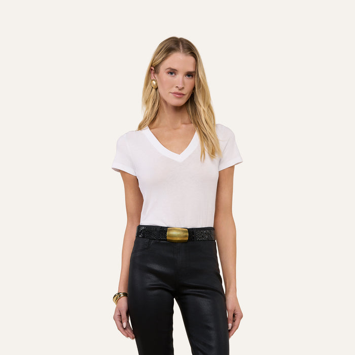 A woman with long blonde hair wears the Becca Cotton V-Neck Tee—crafted from 100 cotton in white—paired with black leather pants featuring a large gold buckle and gold jewelry, standing against a plain light background.