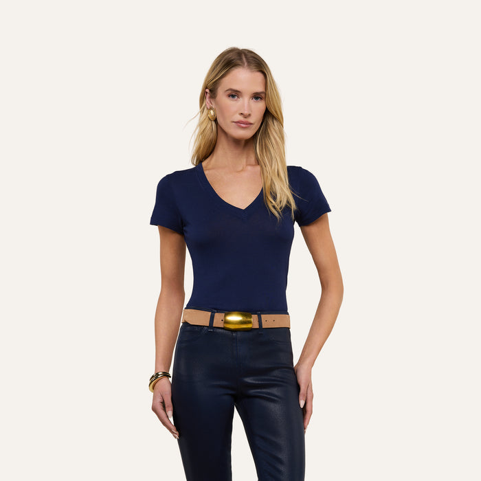 A woman with long blonde hair wears the Becca Cotton V-Neck Tee in navy, pairing it with dark pants, a tan belt featuring a gold buckle, and gold bracelets as she stands against a plain light background.