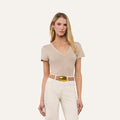 A woman with long blonde hair, wearing the Becca Cotton V-Neck Tee in taupe, white pants, a beige belt with a gold buckle, and pearl earrings stands against a plain light background.