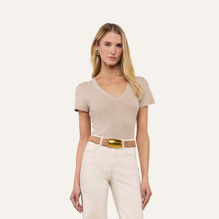 A woman with long blonde hair, wearing the Becca Cotton V-Neck Tee in taupe, white pants, a beige belt with a gold buckle, and pearl earrings stands against a plain light background.