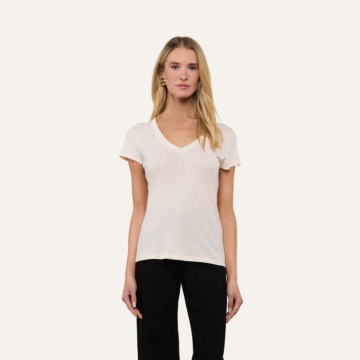 A woman with long blonde hair wears the Becca Cotton V-Neck Tee, a vintage off-white 100 cotton top, paired with black pants. She stands against a plain light background, facing forward with a neutral expression.