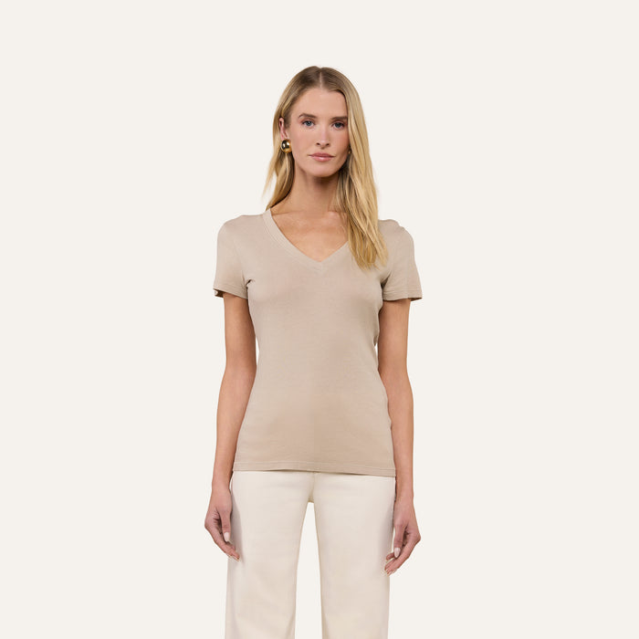 A woman with long blonde hair wears the Becca Cotton V-Neck Tee in a slim fit taupe style, paired with white pants. She stands against a plain light background, facing forward with a neutral expression.
