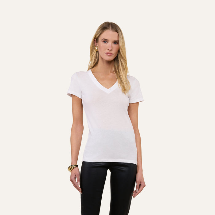 A woman poses against a plain background wearing the Becca Cotton V-Neck Tee, crafted from 100 cotton in white, paired with black pants, gold hoop earrings, and bracelets, her arms relaxed at her sides.
