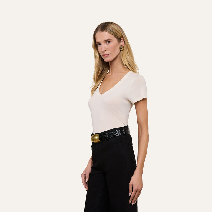 A woman with long blonde hair wears the Becca Cotton V-Neck Tee in a vintage off-white shade, paired with black pants, a black belt featuring a gold buckle, and large round earrings, standing against a plain light background.
