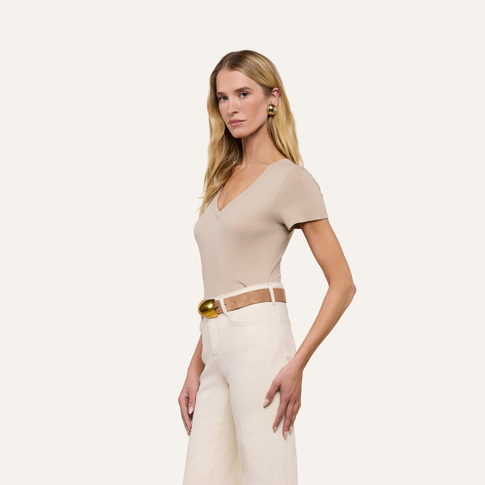 A woman with long blonde hair wears the Becca Cotton V-Neck Tee in taupe, styled with white pants, a tan belt featuring a gold buckle, and earrings, as she stands against a plain light background.