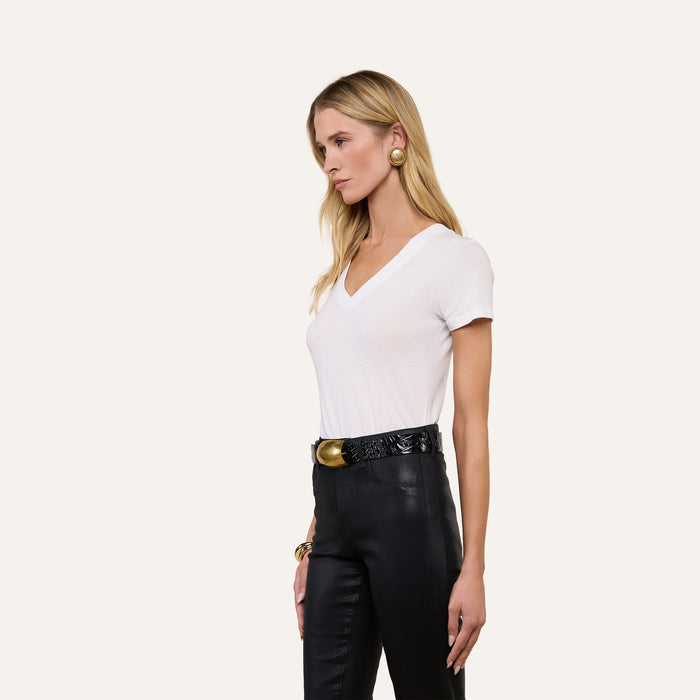 A woman with long blonde hair is wearing the Becca Cotton V-Neck Tee—crafted from 100 cotton—in white, paired with black leather pants, a black belt with a gold buckle, gold earrings, and a bracelet as she stands against a plain light background.