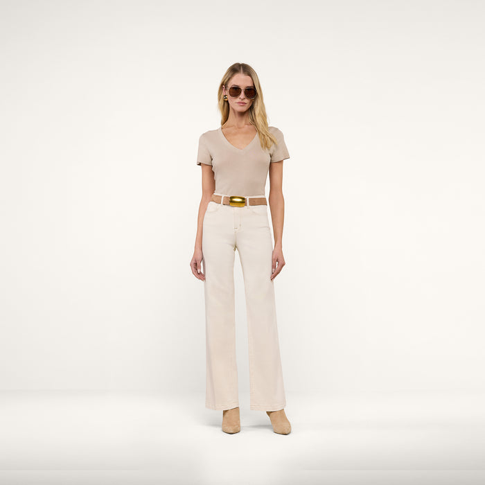 A woman wearing sunglasses and the Becca Cotton V-Neck Tee— a slim fit taupe top— paired with cream wide-leg pants, a white belt with a gold buckle, and beige shoes, standing against a plain white background.