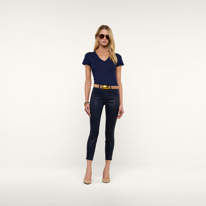 A woman wearing sunglasses, the Becca Cotton V-Neck Tee in navy blue, black fitted pants, a beige belt, gold bracelet, and nude heels stands against a plain white background.
