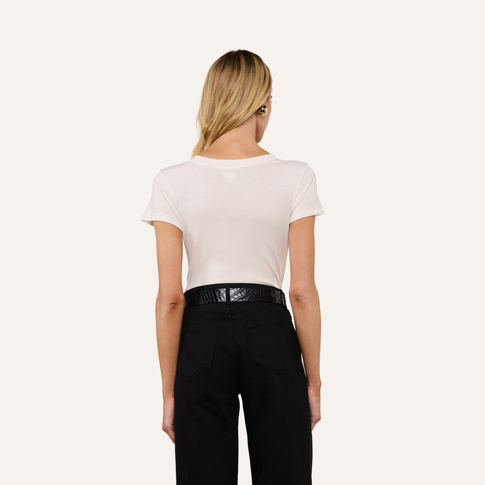 A person with long blonde hair, wearing the Becca Cotton V-Neck Tee in vintage off-white and black pants with a black belt, stands facing away from the camera against a plain background.