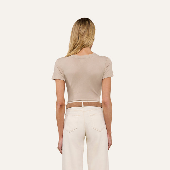 A woman with long blonde hair is shown from behind, wearing the Becca Cotton V-Neck Tee in a slim fit taupe style and high-waisted white pants against a plain, light background.
