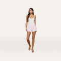 A woman with long brown hair stands against a plain, light background, looking slightly to the side. She wears the Beck Raw-Hem Jean Short in light pink high-rise denim, paired with a white tank top and sandals.