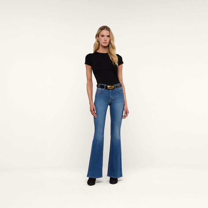 A woman stands facing forward against a plain white background, wearing a black short-sleeve top, the Bell Flare Jean in blue vintage-inspired premium stretch denim, a black belt with a gold buckle, and black shoes.