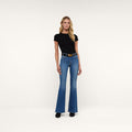 A woman stands facing forward against a plain white background, wearing a black short-sleeve top, the Bell Flare Jean in blue vintage-inspired premium stretch denim, a black belt with a gold buckle, and black shoes.
