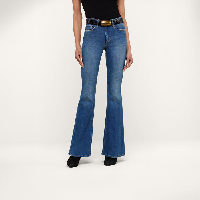 A person wearing the Bell Flare Jean in vintage-inspired blue premium stretch denim, styled with a black sleeveless top, a wide black belt featuring a large buckle, and black shoes, stands against a plain white background.
