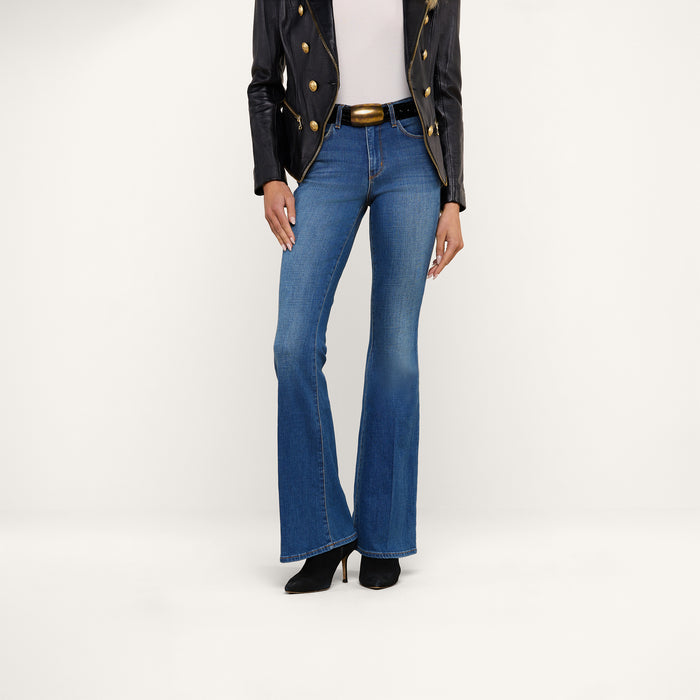 A person wearing the Bell Flare Jean—a pair of blue, vintage-inspired bell-bottom jeans crafted from premium stretch denim—paired with a black jacket, white top, and black heeled boots stands against a plain white background.