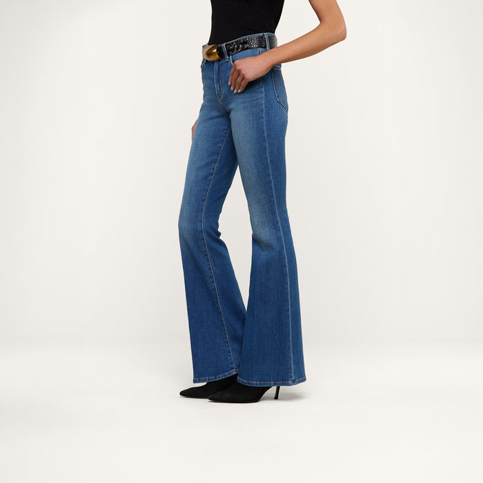 A woman stands against a plain white background wearing the Bell Flare Jean, vintage-inspired blue bell-bottom jeans crafted from premium stretch denim, paired with black pointed-toe heels, a black top, and a black belt featuring a large round gold buckle.