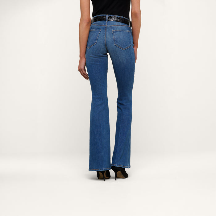 A person stands facing away, wearing the Bell Flare Jean in blue vintage-inspired premium stretch denim, paired with a black top and black heeled shoes against a plain white background.
