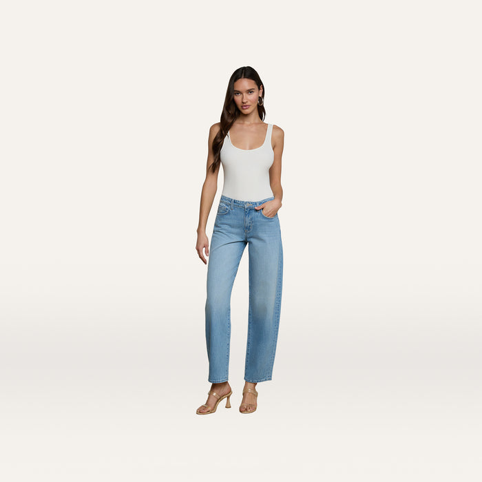 A woman stands against a plain white background wearing a white tank top, the Bex Rodeo Jean in light blue high-waisted barrel leg all-cotton denim, and beige heeled sandals.