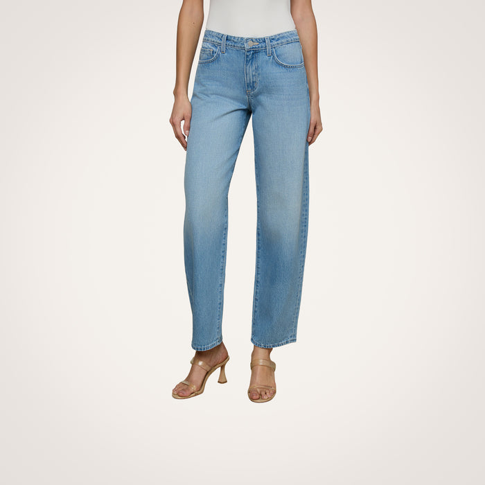 A person wearing the Bex Rodeo Jean in light blue all-cotton denim, paired with a white top and beige heeled sandals, standing against a plain light background.