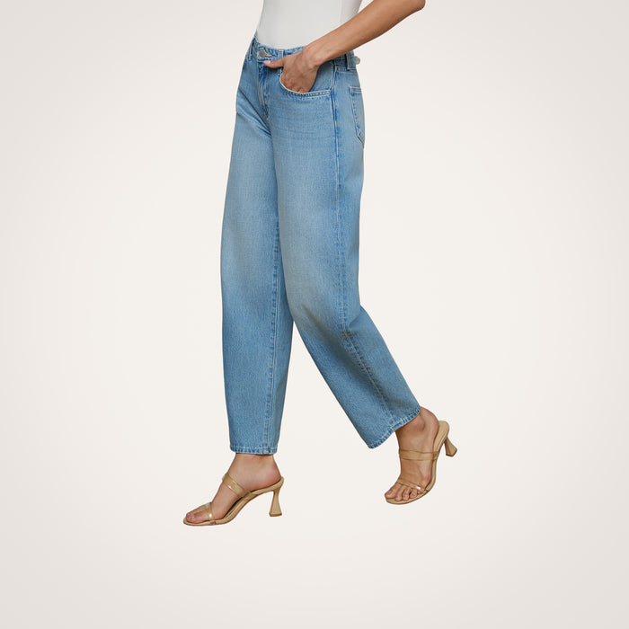 Wearing the Bex Rodeo Jean in light blue, a white top, and beige heeled sandals, a person stands against a plain light background.