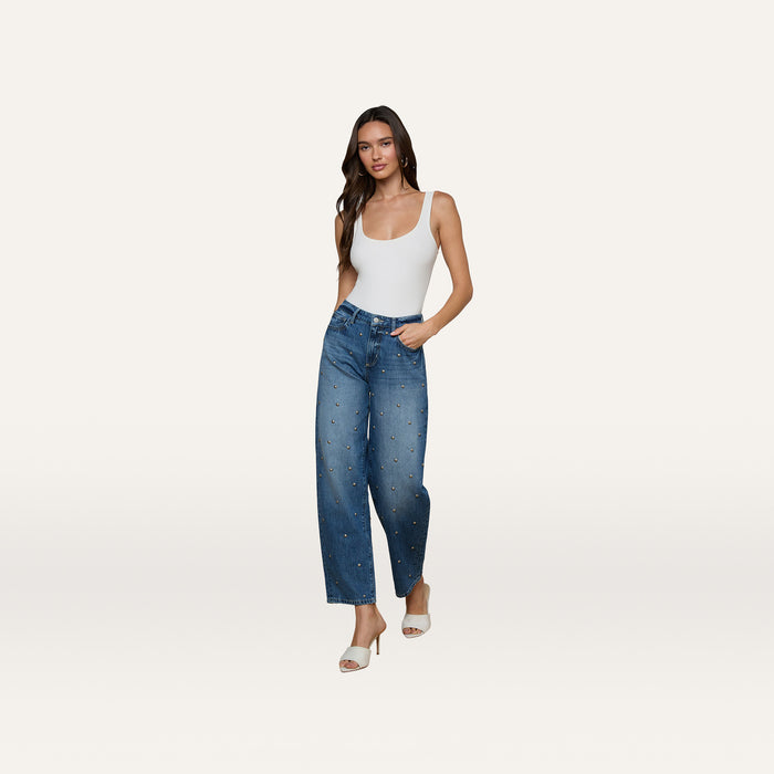A woman stands against a plain white background wearing a white tank top, the Bex Studded Rodeo Jean in blue high-rise with small studded denim embellishments, and white open-toe heels.