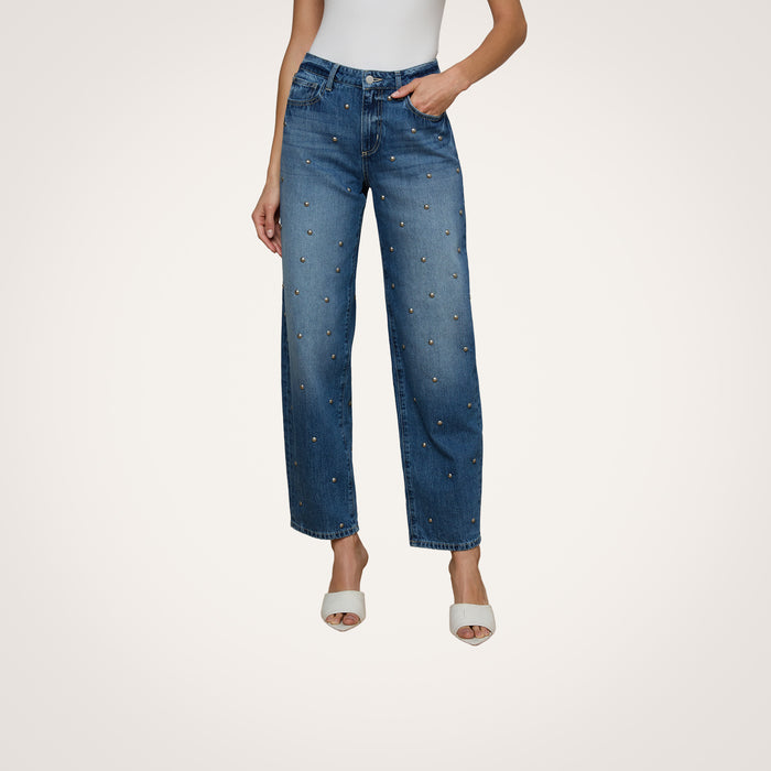A woman wears the Bex Studded Rodeo Jean—high-rise, wide-leg denim featuring small studs—paired with a white sleeveless top and white open-toe heels, standing against a plain background.