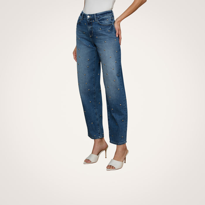 A person is wearing the Bex Studded Rodeo Jean, a pair of high rise blue jeans with small embellishments, styled with a white top and white open-toe heeled sandals, posed against a plain light background.