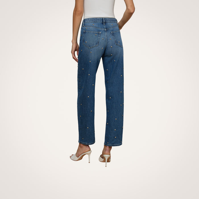 A person wearing a white sleeveless top, the Bex Studded Rodeo Jean in blue with small embellishments, and white high-heeled shoes is shown from the back against a plain background.