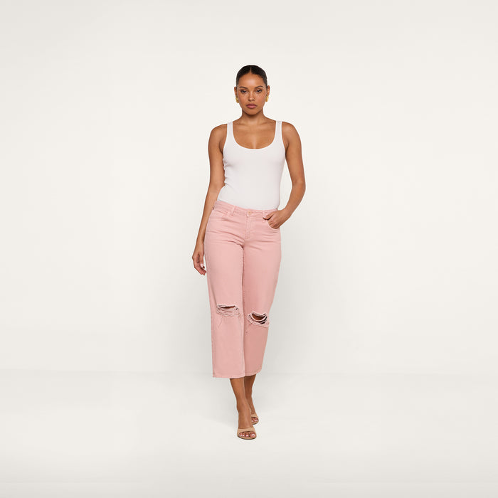 A woman stands against a plain white background wearing a white tank top, the Blaine Cropped Stovepipe Jean in light pink with a straight-leg fit and low rise, and heeled sandals, with one hand in her pocket.