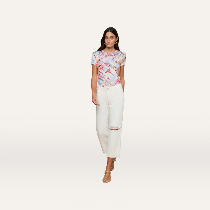 A woman wearing a colorful graphic t-shirt, Blaine Cropped Stovepipe Jeans made from all-cotton denim, and beige heeled sandals stands against a plain white background.