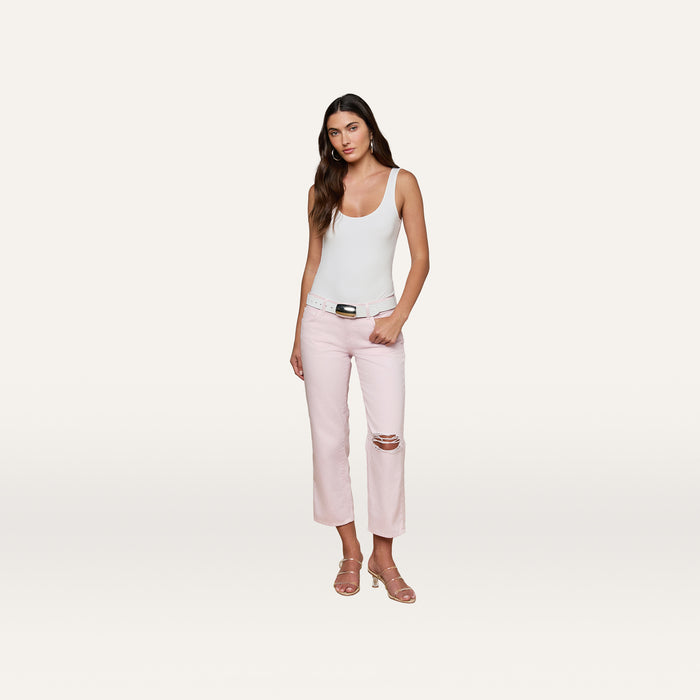 A woman stands against a plain background, wearing a white tank top and the Blaine Cropped Stovepipe Jean in pastel lilac with a belt. She pairs the look with pink sandals, has long brown hair, and maintains a neutral expression.