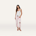 A woman stands against a plain background, wearing a white tank top and the Blaine Cropped Stovepipe Jean in pastel lilac with a belt. She pairs the look with pink sandals, has long brown hair, and maintains a neutral expression.