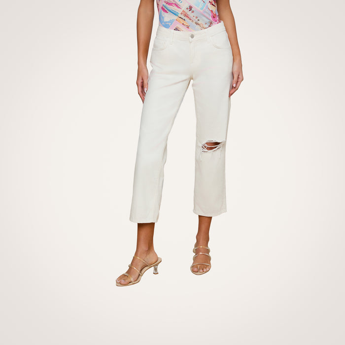 A woman wears the Blaine Cropped Stovepipe Jean in cream-colored, all-cotton denim featuring a rip at one knee, paired with a colorful patterned top and nude heeled sandals, standing against a plain white background.