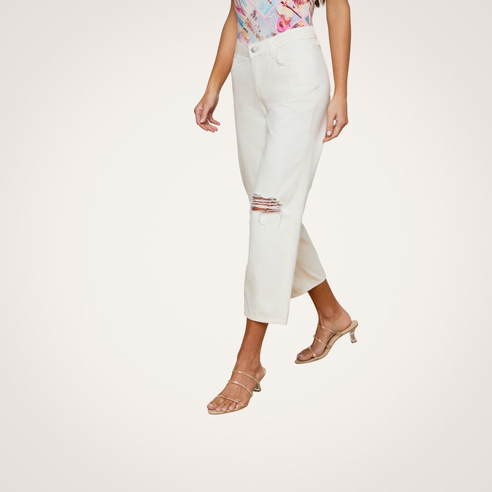 A woman wearing the Blaine Cropped Stovepipe Jean—light-colored, all-cotton denim with a ripped knee—paired with a colorful top and beige heeled sandals, stands against a plain background.