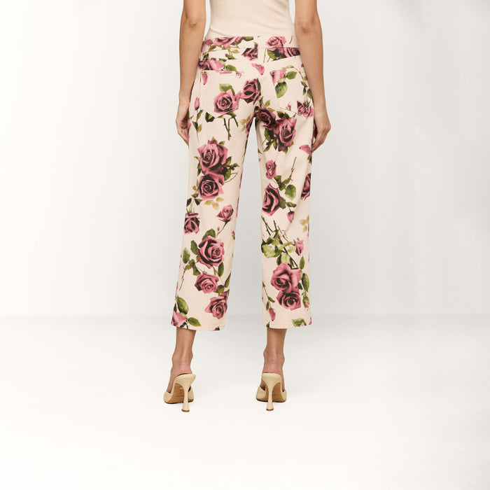 A person stands facing away, wearing the Blaine Cropped Stovepipe Jean in a cream hue with a rose print, paired with a beige top and beige heeled sandals against a plain white background.