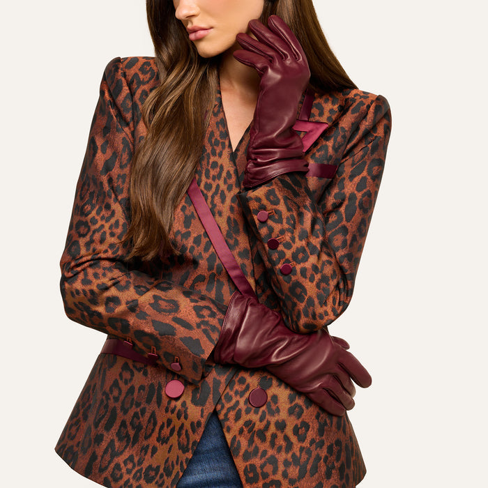 A woman with long brown hair is pictured wearing a leopard print blazer accented with burgundy trim and buttons, her lower face visible as she poses with one arm crossed and the other hand—elegantly gloved in Boheme Gloves crafted from luxury burgundy lambskin leather—resting thoughtfully beneath her chin.