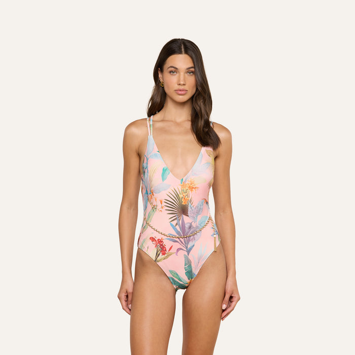 A woman stands facing forward against a plain light background, wearing the Breana Plunge One-Piece Swimsuit featuring a colorful tropical palm and floral print.