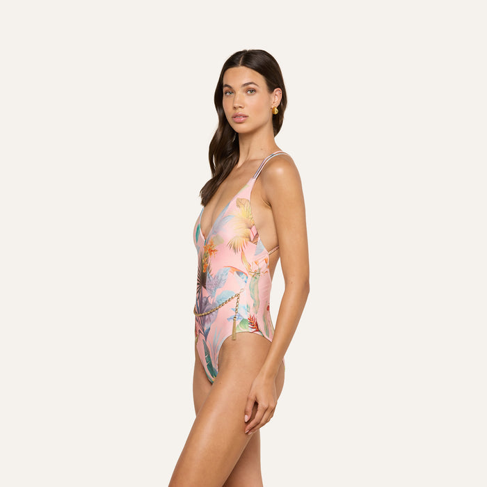 A woman with long brown hair models the Breana Plunge One-Piece Swimsuit—a pink, tropical-print design featuring a crisscross back—shown from the side against a plain light background. She has a neutral expression and wears small hoop earrings.