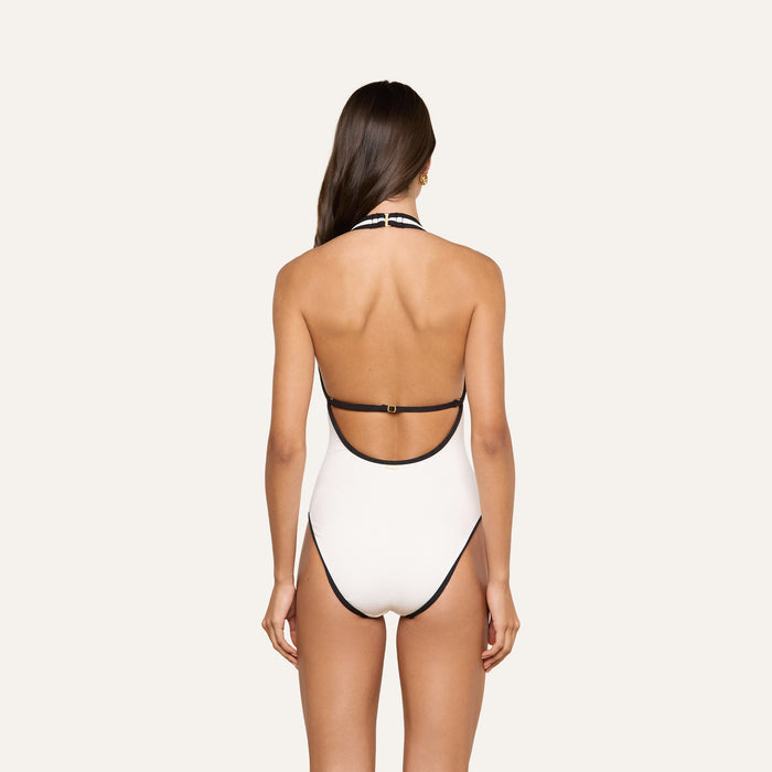 A woman with long brown hair stands facing away, wearing the Breana Plunge One-Piece Swimsuit—UPF 50 white swimwear featuring black trim and an open back—set against a plain light background.