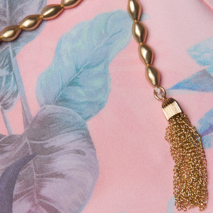 Gold beaded necklace with a chain tassel lying on pink fabric featuring blue and green leafy patterns, reminiscent of the vibrant hues in the Breana Plunge One-Piece Swimsuit.