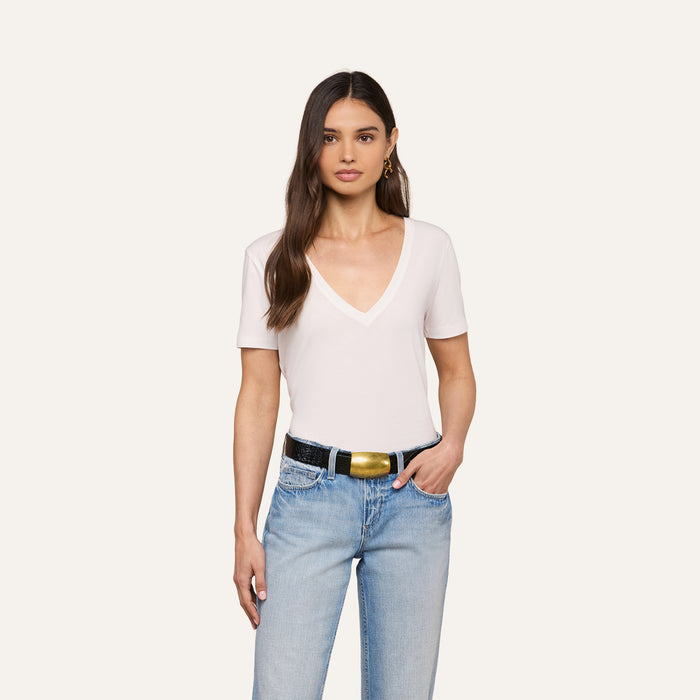 A woman with long brown hair wears a white Brianna Cotton V-Neck Tee, light blue jeans, a black belt with a gold buckle, and gold earrings, standing against a plain light background.