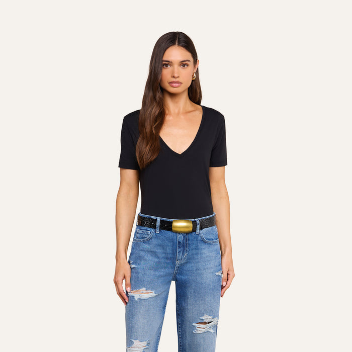 A woman with long brown hair stands against a plain light background wearing a black Brianna Cotton V-Neck Tee made from Supima cotton, paired with blue ripped jeans and a black belt with a gold buckle.