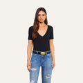 A woman with long brown hair stands against a plain light background wearing a black Brianna Cotton V-Neck Tee made from Supima cotton, paired with blue ripped jeans and a black belt with a gold buckle.