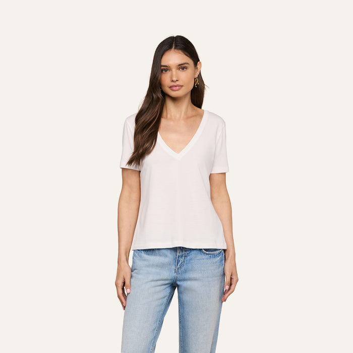A woman with long brown hair wears a white Brianna Cotton V-Neck Tee and light blue jeans, standing against a plain off-white background.