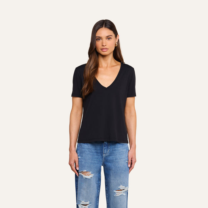 A woman with long brown hair is wearing the Brianna Cotton V-Neck Tee in black, which features a V-neck and short sleeves, paired with ripped blue jeans as she stands facing forward against a plain light background.